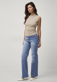 Beige ærmeløs top med rynker ved taljen, parret med lyseblå flared jeans og spidse hvide hæle på en ensfarvet baggrund.