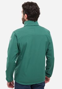 Veste soft-shell verte avec col haut, texture lisse et manches longues. Présente un design ajusté et un logo visible sur la manche gauche.