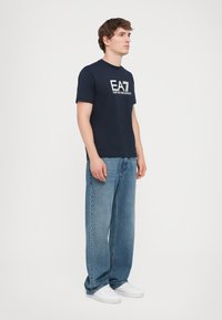 Maglietta blu navy con un logo bianco stampato sul petto, abbinata a jeans azzurri larghi e sneakers bianche.