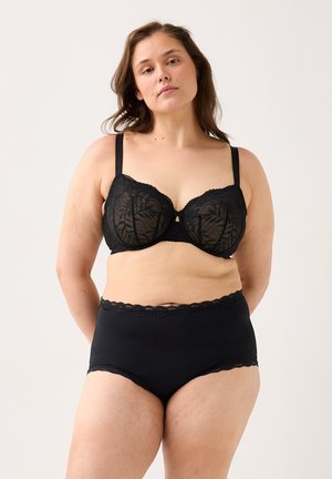 Žena větší velikosti, oblečená v černé krajkové podprsence a černých kalhotkách s vysokým pasem, stojící na jednobarevném světlém pozadí.