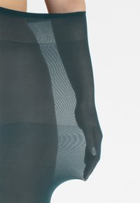 Collant trasparenti color teal con una consistenza liscia e trasparente e una ampia cintura elastica. Il tessuto ha una leggera lucentezza che migliora il design snellente.