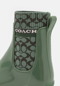 Botas Chelsea de goma verde con calcetín de punto en tono verde oscuro, con una banda blanca con logo y detalles elásticos laterales. Textura lisa en todo el calzado.