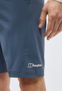 Κοντινό πλάνο του χεριού ενός ατόμου δίπλα σε μπλε σορτς Berghaus με ορατές ραφές και λογότυπο κοντά στο γόνατο.