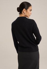 Črn pleteni cardigan s šalom in rebrastim robom, dolgimi rokavi in sproščenim krojem, nosi se čez črno krilo.
