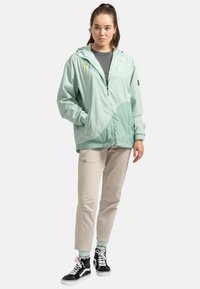 Lichtgroene windbreaker jas met een capuchon, rits aan de voorkant en logo, gecombineerd met beige broek en zwarte hoge sneakers.