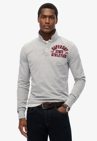 Lichtgrijze langemouwen polo met een knoopkraag, voorzien van rode tekst en logo's op de borst. Gecombineerd met donkere spijkerbroek en eenbruine riem.