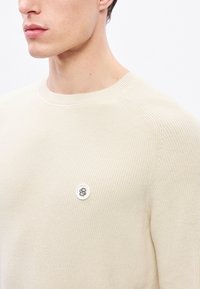 Béžový rebrovaný pletený sveter s okrúhlym výstrihom a bielym gombíkom s emblémom v tvare štýlizovaného „S“ na ľavej strane hrudníka.