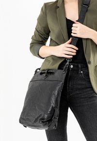 Borsa a spalla in pelle nera con finitura testurizzata, manico superiore, tracolla regolabile e patta frontale. Indossata con un blazer verde oliva e jeans.