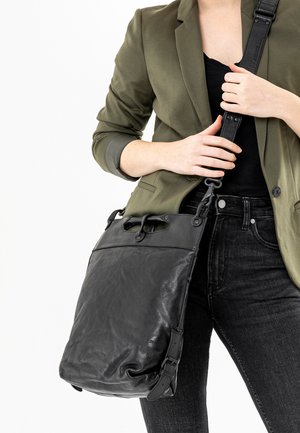 Schwarze Leder-Schultertasche mit strukturiertem Finish, oberem Griff, verstellbarem Riemen und vorderer Klappe. Getragen zu einem olivgrünen Blazer und Jeans.
