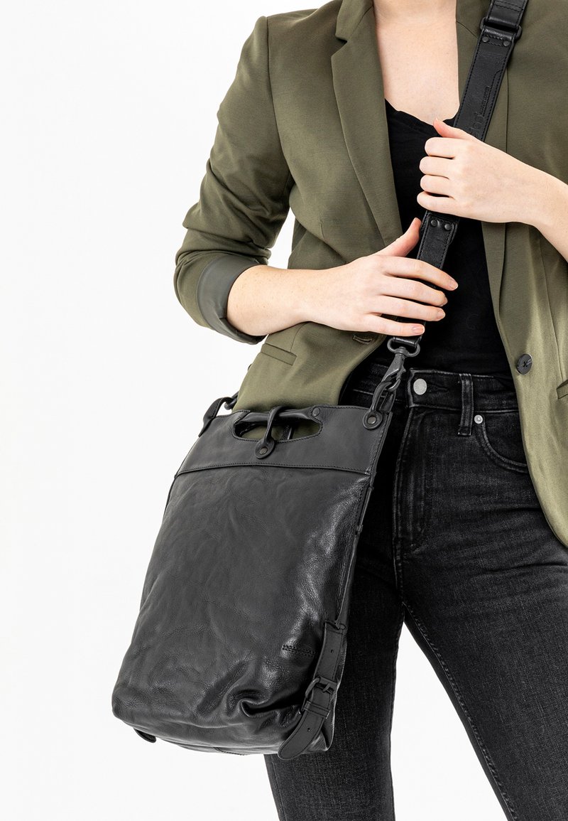 Borsa a spalla in pelle nera con finitura testurizzata, manico superiore, tracolla regolabile e patta frontale. Indossata con un blazer verde oliva e jeans.