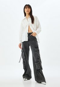 Camicia bianca con bottoni e borchie decorative, abbinata a pantaloni cargo neri oversize con numerose tasche. Modello che indossa scarpe metalliche.