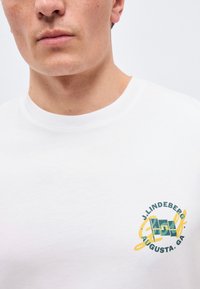 Moški, ki nosi belo majico s kratkimi rokavi z zeleno in rumeno nalepko »J. Lindeberg Augusta, GA« na levi prsni strani.