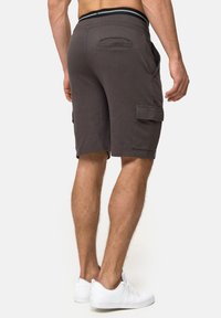 Mand iført mørkegrå cargo-shorts og hvide sneakers, står med ryggen og benene synlige mod en ensfarvet lys baggrund.