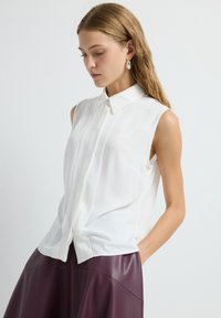 Weißes ärmelloses Bluse mit spitzem Kragen, ausgestattet mit einer Knopfleiste und glatter Textur, kombiniert mit einem burgunderroten Lederskirt.