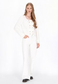 Witte geribbelde cardigan met ronde halslijn, gouden knopen en lange mouwen, gecombineerd met witte flared jeans en zwarte schoenen.