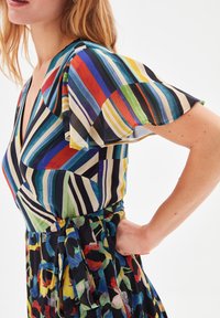 Robe multicolore avec des rayures verticales et horizontales, manches courtes, et un motif floral sur un fond sombre. Détail de taille nouée visible.