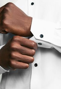 Chemise blanche avec une finition texturée, ornée de boutons noirs sur le poignet. Gros plan sur une main attachant le poignet.
