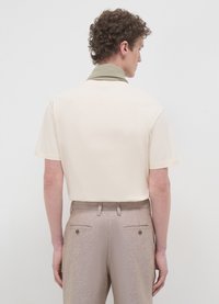 Camicia crema a maniche corte con colletto in olivo texture, abbinata a pantaloni marrone chiaro con due tasche posteriori e un leggero lucido.