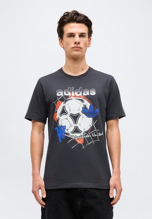 Jeune homme portant un t-shirt Adidas gris foncé avec un motif de ballon de football, logo bleu de Manchester United et pantalon noir, debout sur fond blanc.