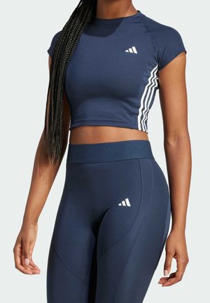Vrouw die een marineblauwe Adidas crop top en een high-waisted legging met witte strepen en logo’s draagt, staat rechtop met één arm langs haar zijde.