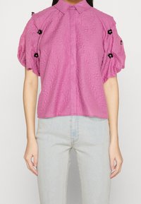 Blusa rosa con texture, colletto e maniche a sbuffo, con dettagli decorativi neri sulle spalle.