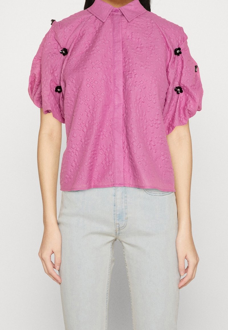 Blusa rosa con texture, colletto e maniche a sbuffo, con dettagli decorativi neri sulle spalle.