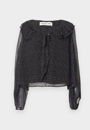 Kobieta stojąca w przezroczystej czarnej bluzce z falbankami i czarnych spodniach z haftowanymi rozcięciami przy kostkach oraz czarnych szpiczastych butach.