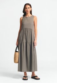 Nesuknijos pilka maksi suknelė su sluoksnių dizainu, minkšta tekstūra ir apvaliu iškirpimu; derinama su pintine kelių maišeliu ir juodais butais sandalais.