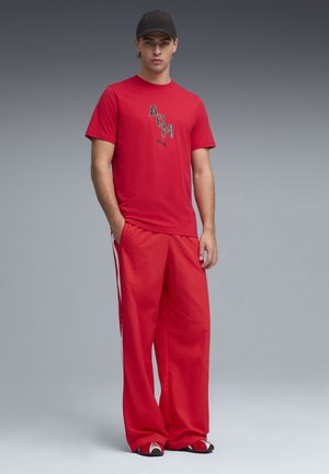 Młody mężczyzna w czerwonej koszulce ACM Puma, szerokich czerwonych spodniach z białymi bocznymi paskami, czapce z daszkiem i czerwono-białych trampkach stoi z rękami w kieszeniach.
