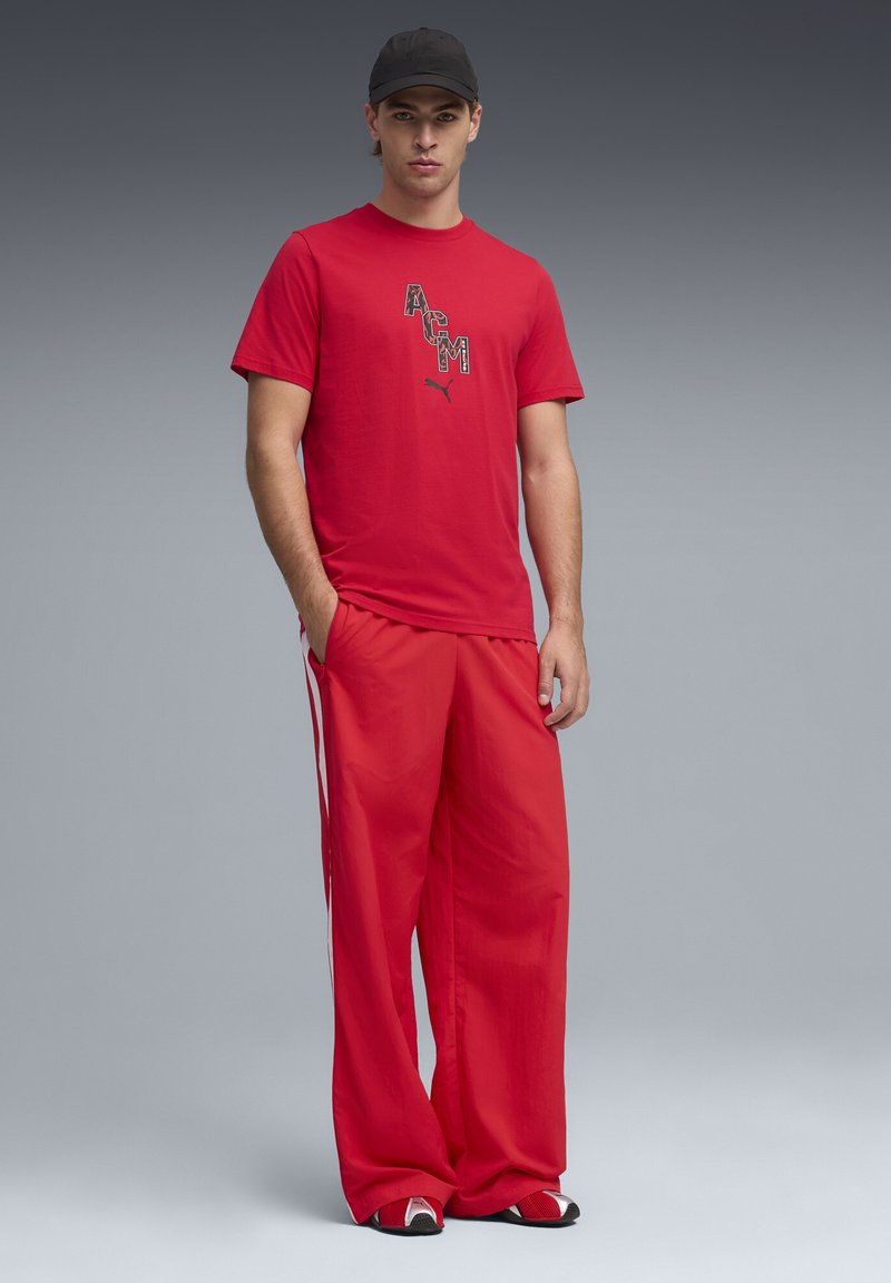 Unge mand iført en rød ACM Puma T-shirt, røde viddebukser med hvide sidestriber, sort kasket og røde-hvide sneakers står med hænderne i lommerne.