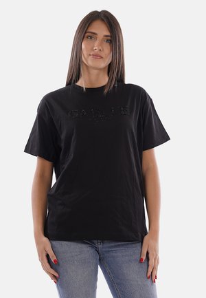 Giovane donna con capelli castani lisci che indossa una maglietta nera con una scritta sottile "GAELLE" e jeans blu, in piedi davanti a uno sfondo semplice.
