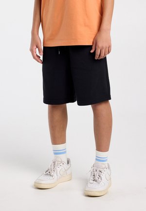 Persona vistiendo pantalones cortos negros hasta la rodilla, camiseta naranja, calcetas blancas con rayas azules y zapatillas blancas, de pie sobre un suelo gris claro.