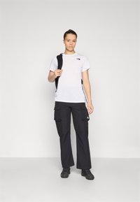T-shirt bianco con logo nero, pantaloni cargo neri con tasche e scarpe nere, indossati da una persona con i capelli raccolti, su uno sfondo neutro.