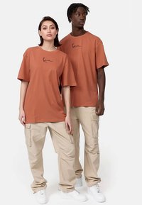 Dwóch modeli noszących oversize'owe, rdzawe bawełniane koszulki z czarnymi akcentami logo, w połączeniu z beżowymi spodniami cargo i białymi trampkami.