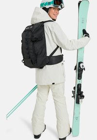 Weiße Outerwear mit schwarzen Akzenten, einen schwarzen Rucksack und mintgrüne Skier. Enthält eine Skibrille und Handschuhe, mit einem strukturierten Taschendesign.