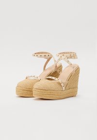 Sandalias de cuña con un upper tejido beige texturizado, tiras crema con tachuelas doradas y una suela media de yute trenzado para mayor altura.