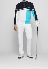 Golfoutfit med en marinblå, vit och turkos zip-up topp med färgblockering, i kombination med vita byxor och vita skor med gröna detaljer.