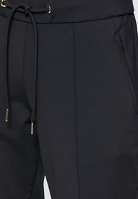 Zwarte sportbroek met een trekkoord in de taille, gouden hardware, een zijzak en een gladde, gestructureerde stof.