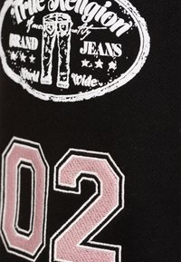 Tissu noir avec un logo blanc présentant des graphismes de style denim, rehaussé d'un "02" brodé en rose avec des détails de bordure blanche.