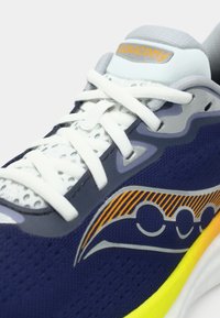 Detailný záber na námorníckou modrú behaciu obuv Saucony s bielymi šnúrkami, šedými akcentmi a logom so žltým pruhom nad žltou podrážkou.