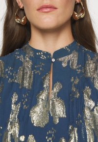 Top azul con patrones florales metálicos dorados, un escote alto con un pequeño corte en forma de llave y un cierre de botón único en el cuello.