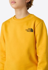 Gul genser laget av mykt stoff med ribbet hals. Har en svart logo på venstre bryst og lange ermer. Glatt tekstur.