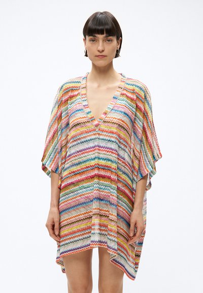 Femme aux cheveux courts et foncés, portant une robe ample et colorée à motif en zigzag, avec un col en V et des manches aux coudes.