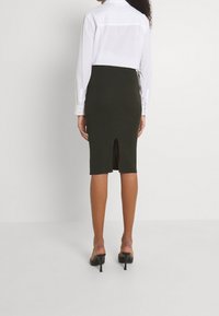 Olivgrön pennkjol med slits bak, tillverkad i stretchigt tyg. Kombinerad med en vit skjorta med knappar och svarta klacksandaler.