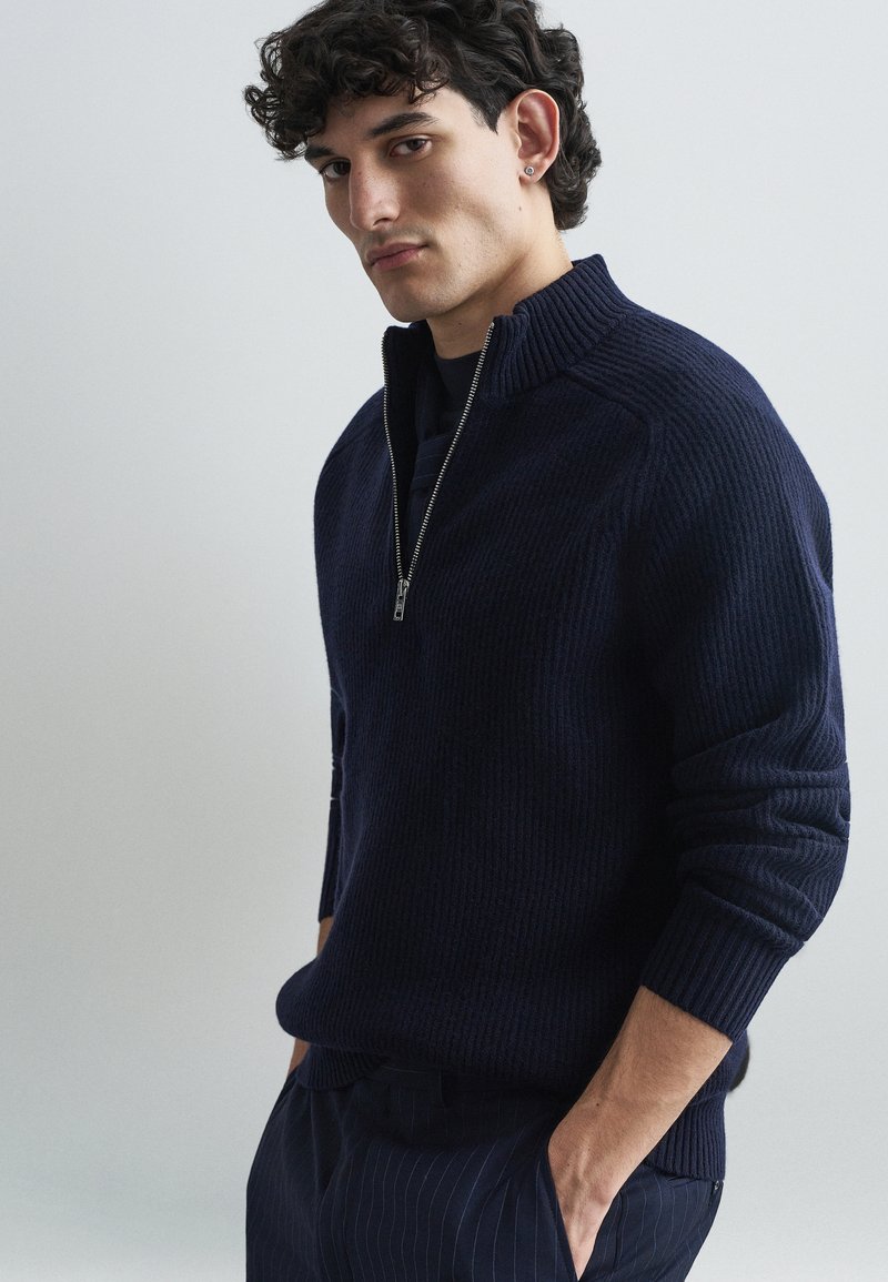 Navy ribbed knit sweater with a half-zip collar, featuring long sleeves and a relaxed fit, paired with pinstriped pants.