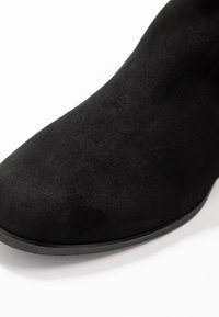 Bottine en suède noir avec une finition lisse, un bout pointu et une semelle plates noire. Pas de quincaillerie ou d'accents visibles.