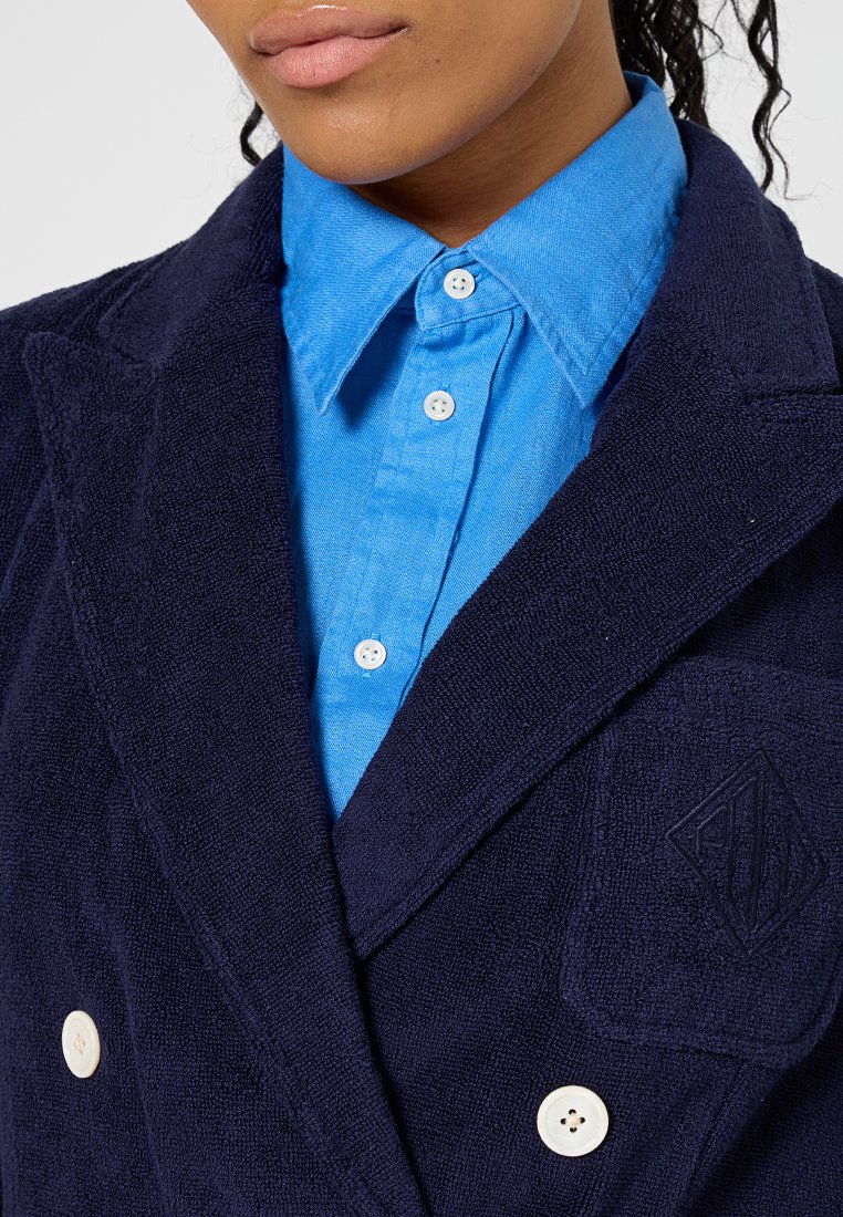 Granatowa marynarka dwurzędowa z froty, z białymi guzikami, o teksturowanym wykończeniu i małym, wytłoczonym logo na kieszeni. Jasnoniebieska koszula pod spodem.
