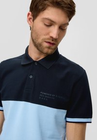 Granatowa i jasnoniebieska koszulka polo z dwukolorowym wzorem, teksturowanym materiałem oraz haftowanym logo na piersi. Klasyczny kołnierzyk z guzikami.