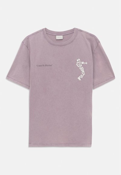 Lavendelfarbenes kurzärmliges T-Shirt mit dem Text "United By Rhythm" auf der linken Brust und einer Grafik eines Trompeters, die aus dem Text "FILMMAKER" besteht, auf der rechten Brust.