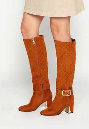 Bottes en daim matelassé orange rouille mi-mollet avec talons blocs, fermeture éclair latérale et détails de boucle dorée, portées avec une jupe beige.